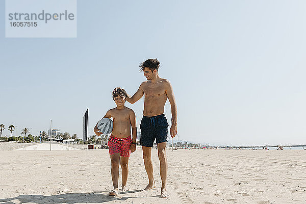 Hemdloser junger Mann mit seinem Neffen bei einem Strandspaziergang gegen den klaren Himmel