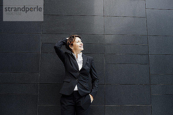 Lächelnde weibliche Fachkraft mit Hand im Haar vor einem modernen Gebäude stehend
