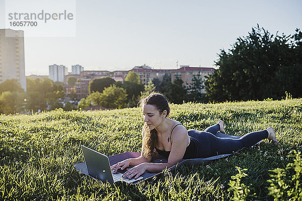 Sportliche junge Frau  die einen Laptop benutzt  während sie auf einer Übungsmatte im Stadtpark liegt