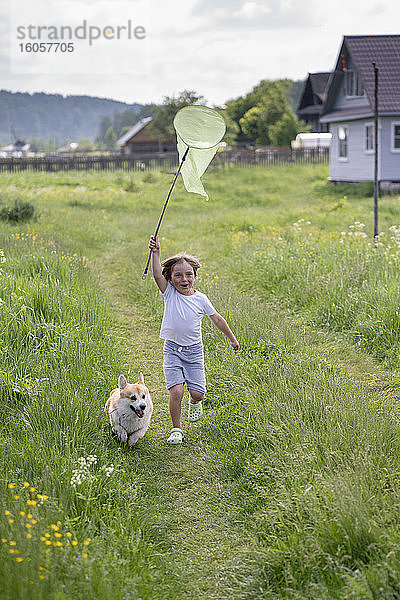 Verspielter Junge  der ein Schmetterlingsnetz hält und mit seinem Hund auf einer Wiese läuft