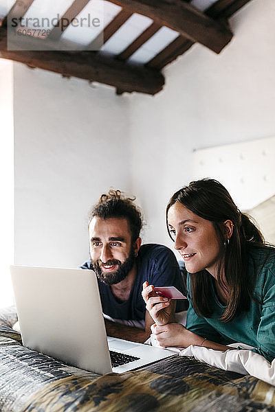 Glückliches Paar liegt auf dem Bett und kauft online ein