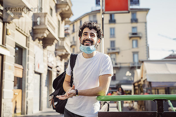 Lächelnder junger Mann mit Gesichtsmaske  der wegschaut  während er an einem Geländer in der Stadt steht