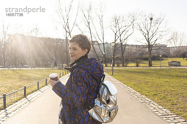 Porträt einer lächelnden Frau mit Rucksack und Kaffee zum Mitnehmen in einem Park  Berlin  Deutschland