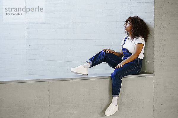 Nachdenkliche junge Frau mit Afro-Haar sitzt auf einer Stützmauer