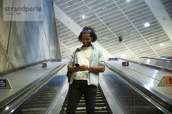 Junger trendiger Mann lächelt  während er sein Smartphone auf einer Rolltreppe in einer U-Bahn-Station benutzt