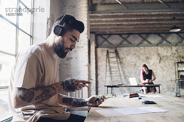 Kreativer junger Geschäftsmann mit Kopfhörern und Smartphone im Loftbüro
