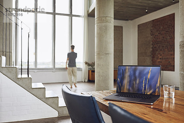Laptop auf Holztisch in einer Loftwohnung mit Mann am Fenster im Hintergrund