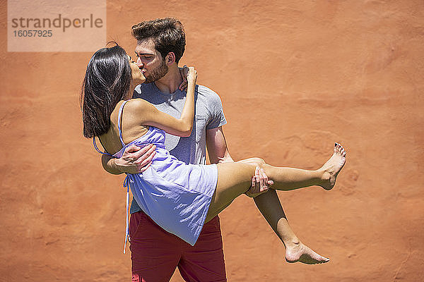 Junger Mann küsst  während er eine Frau gegen eine braune Wand im Hinterhof trägt  während eines sonnigen Tages
