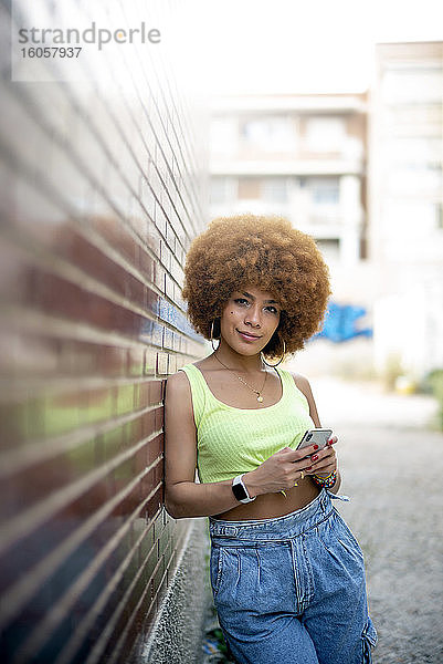 Mittlere erwachsene Frau mit Afro-Haar  die ein Smartphone benutzt  während sie sich an eine Mauer in der Stadt lehnt