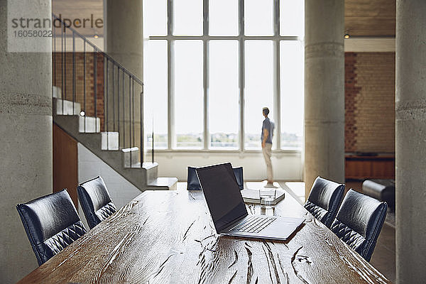 Laptop auf Holztisch in einer Loftwohnung mit Mann am Fenster im Hintergrund