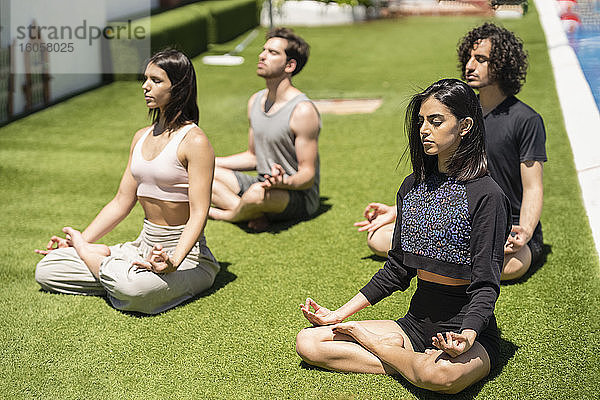 Junge Mala und weibliche Freunde meditieren während sie Yoga auf dem Rasen im Hinterhof bei sonnigem Wetter üben