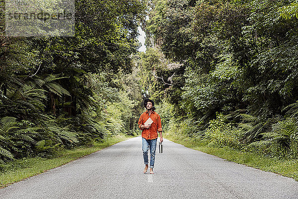 Nachdenklicher junger Mann  der eine Wasserflasche und ein Buch hält  während er auf einer Straße im Wald spazieren geht