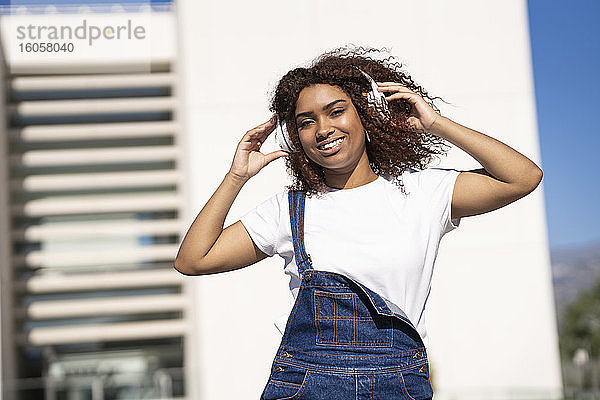 Lächelnde Afro-Frau  die Musik über Kopfhörer hört  während sie an einem Gebäude steht