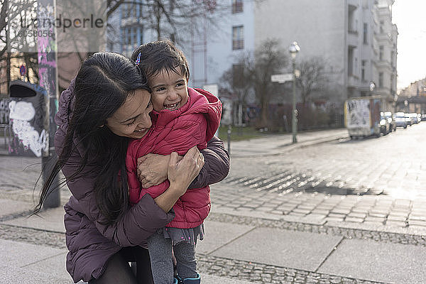 Glückliche Frau  die ihre Tochter umarmt  während sie auf einem Fußweg in der Stadt kniet