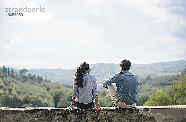 Paar  das die Aussicht betrachtet  während es auf einer Stützmauer gegen den Himmel an einem sonnigen Tag sitzt