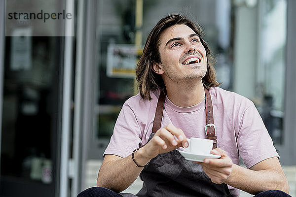 Männlicher Besitzer hält lachend eine Kaffeetasse  während er vor einem Café sitzt