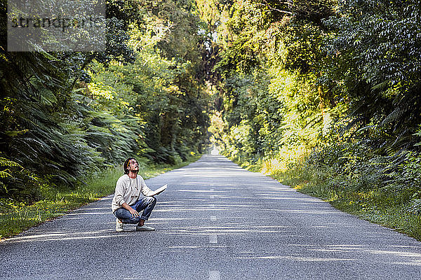Junger Mann mit Tagebuch auf der Landstraße inmitten von Bäumen hockend