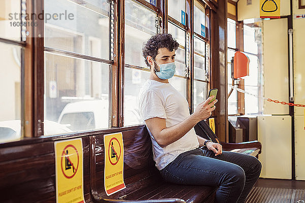 Junger Mann mit Gesichtsmaske  der in der Straßenbahn sitzt und sein Smartphone benutzt