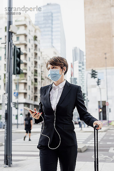 Geschäftsfrau mit Maske  die ein Smartphone hält  während sie mit einem Koffer in der Stadt steht