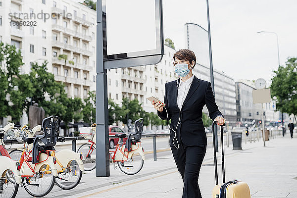 Geschäftsfrau mit Maske  die Musik hört  während sie mit einem Koffer auf dem Gehweg in der Stadt spazieren geht