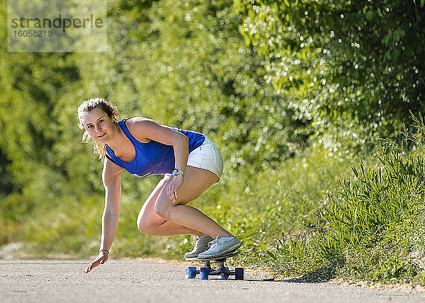 Schöne junge Frau skateboarding auf der Straße von Pflanzen während sonnigen Tag