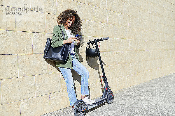 Glückliche Frau  die ihr Smartphone benutzt  während sie mit einem elektrischen Tretroller auf dem Bürgersteig steht