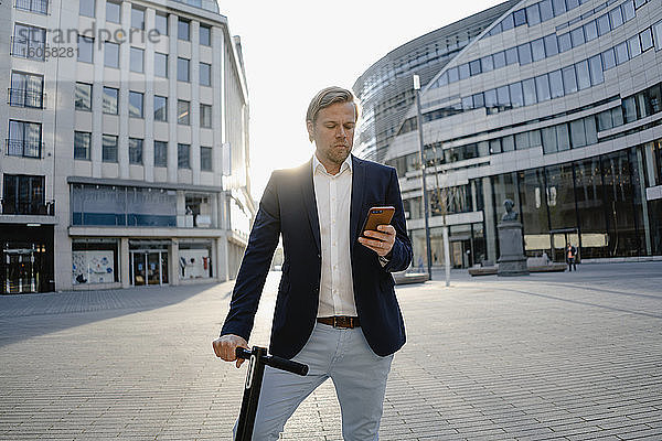 Geschäftsmann mit Kick-Scooter mit Smartphone in der Stadt bei Sonnenuntergang