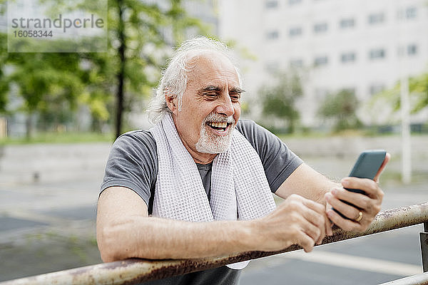 Nahaufnahme eines fröhlichen älteren Mannes  der sein Smartphone benutzt  während er an einem Geländer steht