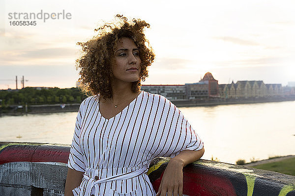 Nachdenkliche Frau mit lockigem Haar steht am Geländer gegen den Fluss in der Stadt bei Sonnenuntergang