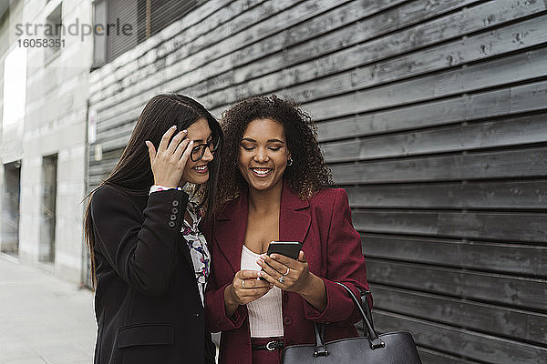 Lächelnde junge multiethnische Geschäftskollegen  die sich ein Smartphone an einer Wand in der Stadt teilen