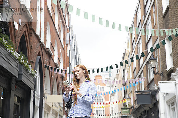 Lächelnde junge rothaarige Frau  die ein Smartphone benutzt  während sie inmitten von Gebäuden in der Stadt steht