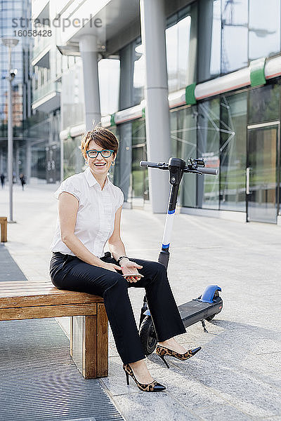 Lächelnde Geschäftsfrau mit elektrischem Tretroller auf dem Sitz in der Stadt