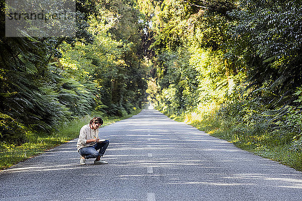 Junger Mann schreibt in Tagebuch  während er auf einer Landstraße inmitten von Bäumen hockt