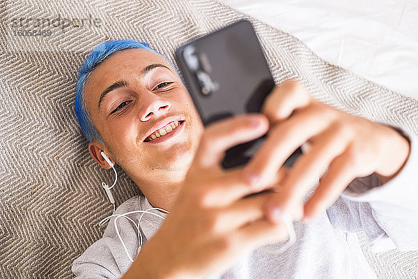 Junger Mann mit blauen Haaren  der auf dem Bett liegend sein Smartphone benutzt und Musik hört