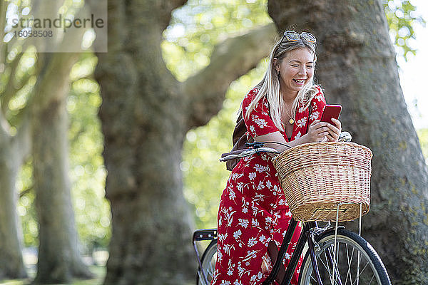 Fröhliche Frau benutzt ihr Smartphone  während sie sich in einem öffentlichen Park auf ein Fahrrad stützt