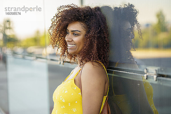 Nahaufnahme einer lächelnden jungen Frau mit Afro-Haar  die an einer Wand steht und wegschaut