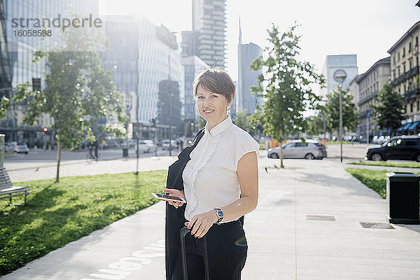 Lächelnde Geschäftsfrau mit Smartphone auf dem Bürgersteig in der Stadt an einem sonnigen Tag