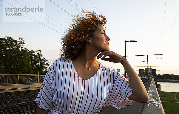 Nachdenkliche Frau mit lockigem Haar steht am Geländer gegen den Himmel bei Sonnenuntergang