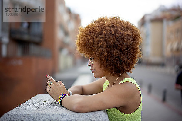 Mittlere erwachsene Frau mit Afro-Haar  die ein Smartphone benutzt  während sie an einer Stützmauer steht