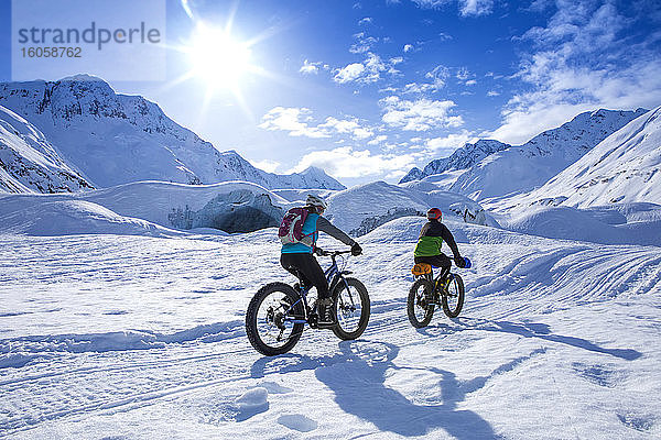 Zwei fette Frauen radeln an einem sonnigen Wintertag vor dem Skookum-Gletscher  Chugach National Forest  Alaska  und pumpen mit der Faust  während sie vorbeifährt; Süd-Zentral-Alaska; Alaska  Vereinigte Staaten von Amerika