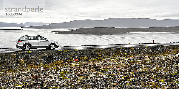 Ein Fahrzeug  das auf einer Straße am Wasser geparkt ist  mit Blick auf den Fjord und silhouettierte Hügel unter bedecktem Himmel; Sudavik  Westfjorde  Island