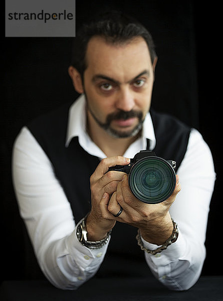 Porträt eines männlichen Fotografen  der mit seiner Kamera vor einem schwarzen Hintergrund steht  Fokus auf die Kamera im Vordergrund; Studio
