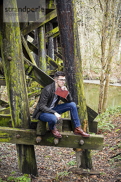 Junger Mann sitzt auf einer moosbewachsenen Gerüstbrücke neben einem Teich mit einem Buch in ruhiger Lage; Bothell  Washington  Vereinigte Staaten von Amerika