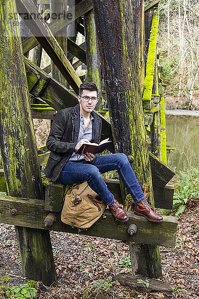 Junger Mann sitzt auf einer moosbewachsenen Bockbrücke neben einem Teich und liest in ruhiger Lage aus einem Buch; Bothell  Washington  Vereinigte Staaten von Amerika