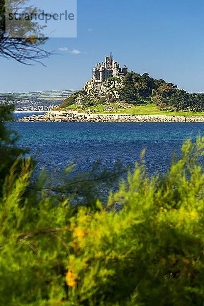 Die Burg St. Michael thront auf einem felsigen Hügel entlang einer Küstenlinie  umgeben von Bäumen mit blauem Himmel  eingerahmt von Laub im Vordergrund; Grafschaft Cornwall  England