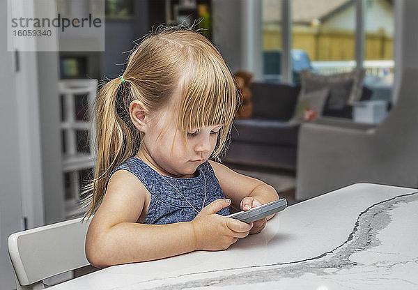 Ein junges Mädchen  das mit ihrem Smartphone spielt  während es an einem Tisch in ihrer Küche sitzt: Edmonton  Alberta  Kanada