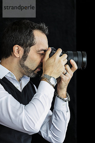 Porträt eines männlichen Fotografen  der vor einem schwarzen Hintergrund steht und seine Kamera benutzt; Studio