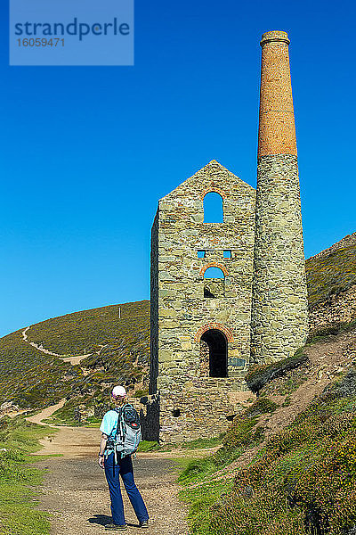 Weibliche Wanderin auf Schotterweg mit Steingebäude und Schornstein einer alten Zinnmine und blauem Himmel; Grafschaft Cornwall  England