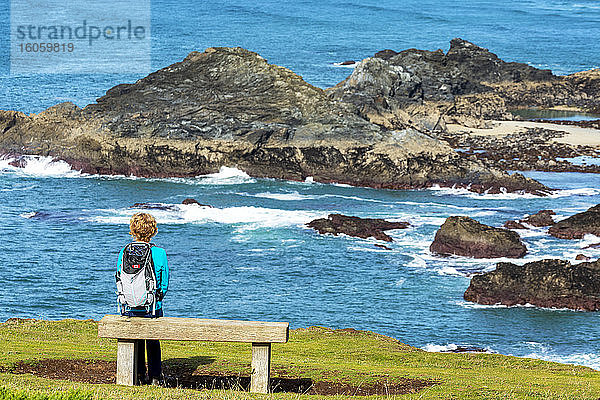 Eine Wanderin sitzt auf einer Holzbank mit Blick auf die felsige Küste; Grafschaft Cornwall  England