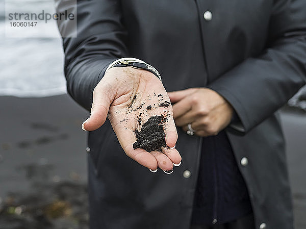 Eine Frau steht mit schwarzem Sand in der Hand; Island